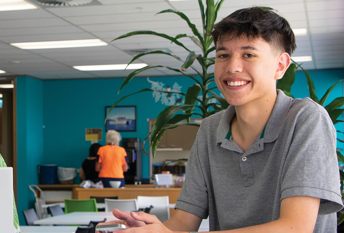 A young man sitting in a room with teal blue wall in the background.