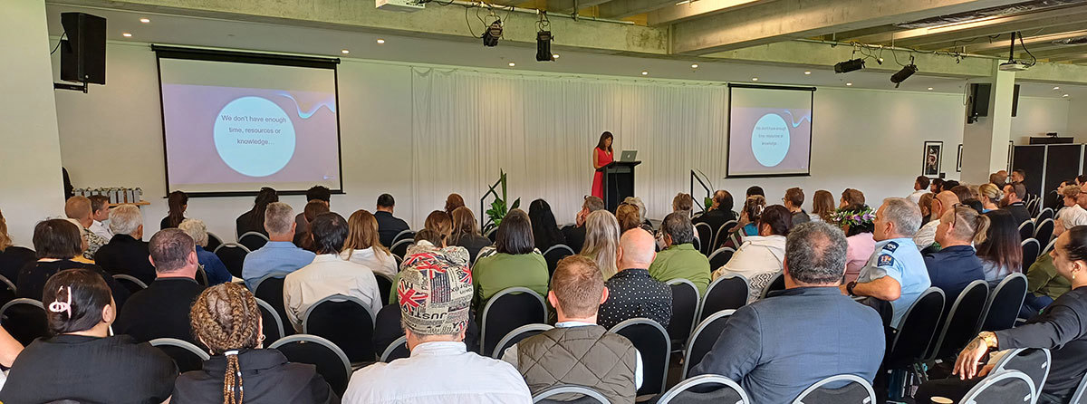 Many people seated in a room watching and listening to a woman presenting on a stage.