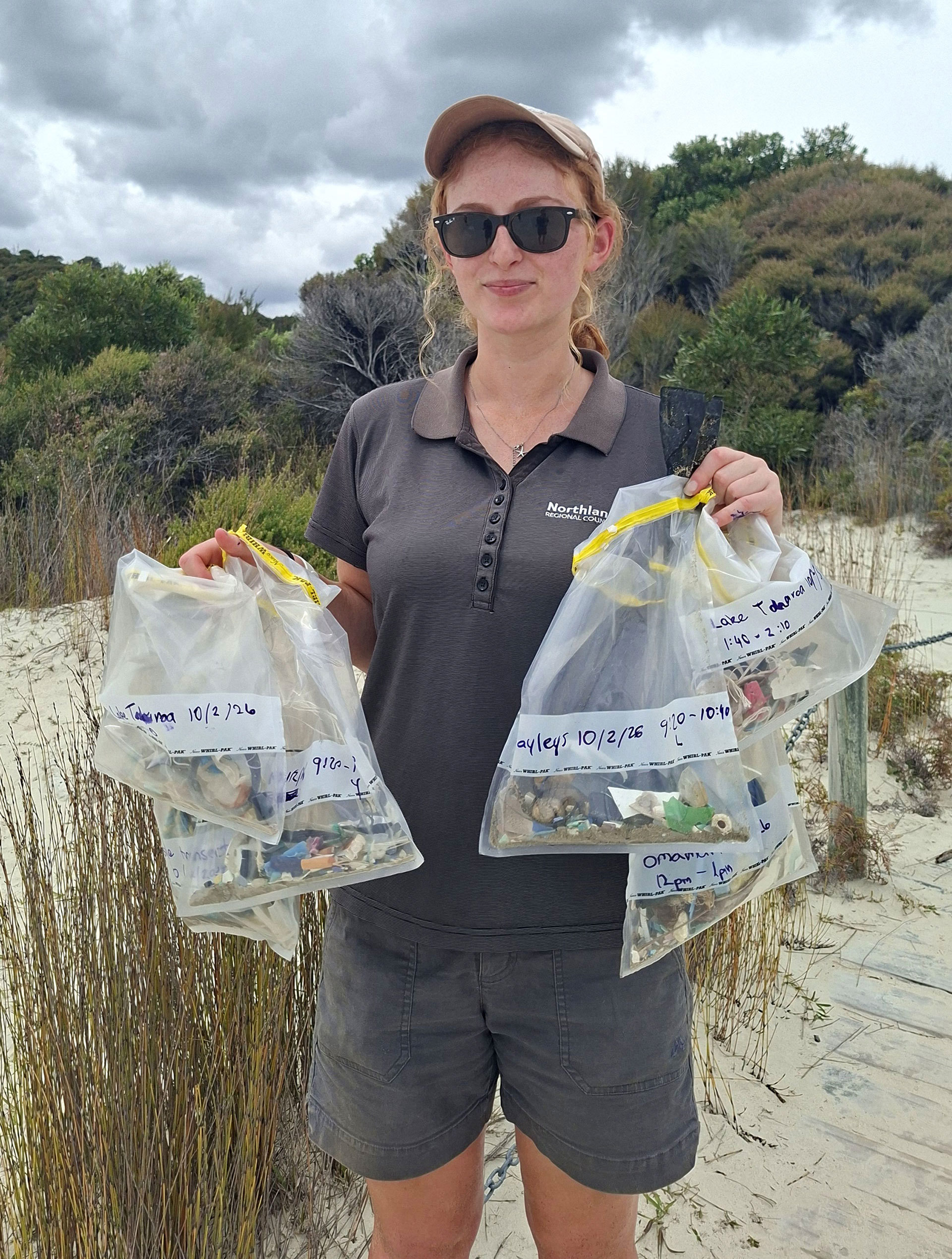 Woman wearing a cap and sunglasses holding several plastic bags containing litter collected from beaches.