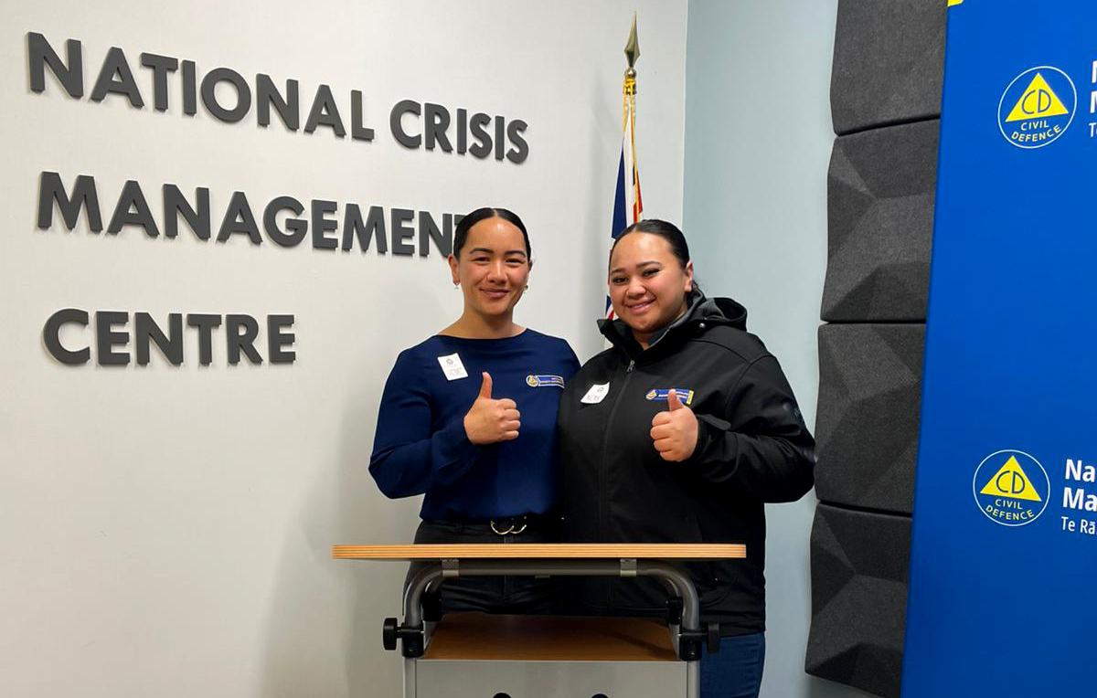 Two women giving a thumbs up behind a podium with National Crisis Management Centre on the wall behind.