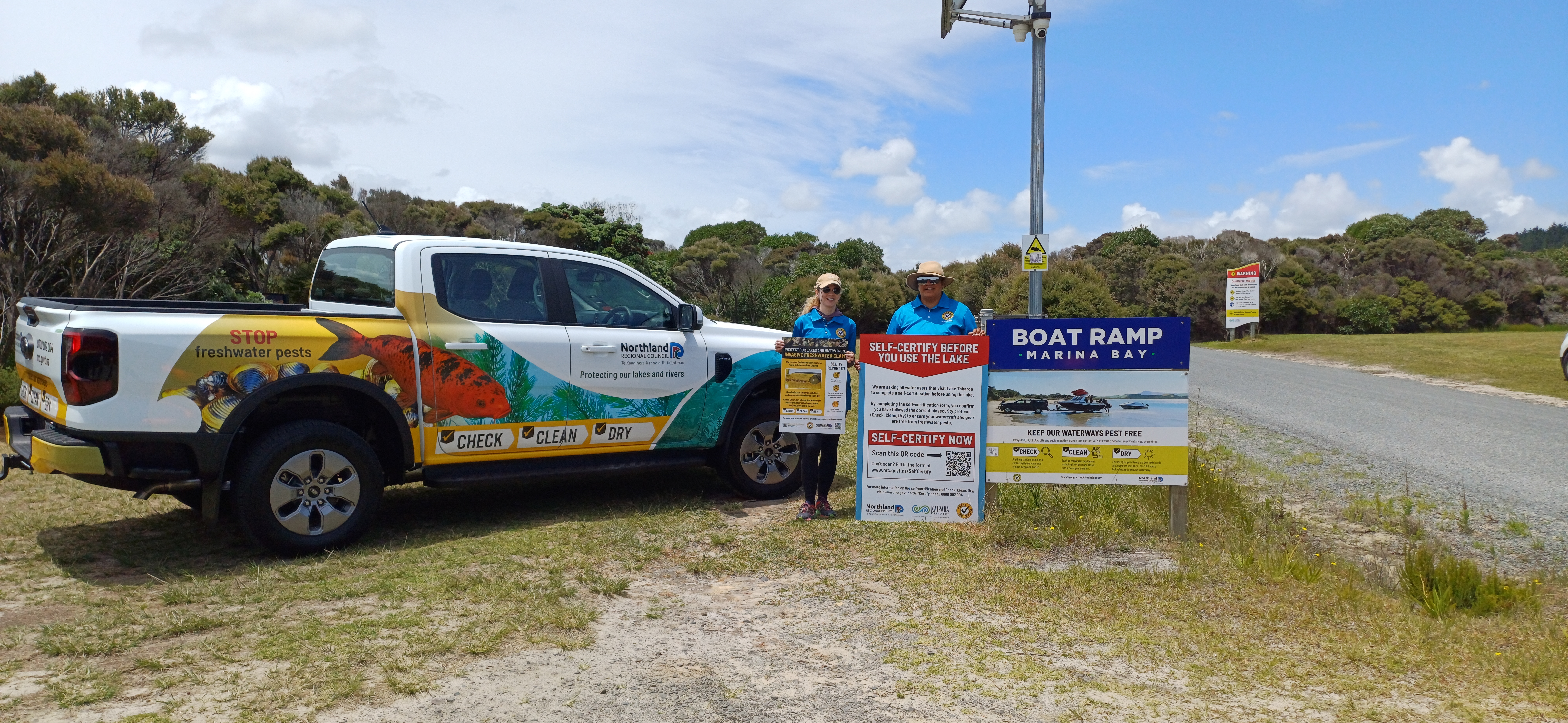 A white pickup truck with colorful biosecurity-themed graphics is parked on a grassy area beside a gravel road. Two people wearing matching blue uniforms and hats stand next to the truck, holding and standing beside large informational signs about “Check, Clean, Dry” and self‑certification for freshwater pest prevention. To the right of them is a prominent “Boat Ramp – Marina Bay” sign with additional safety and biosecurity information. Bushes and trees fill the background under a partly cloudy sky.