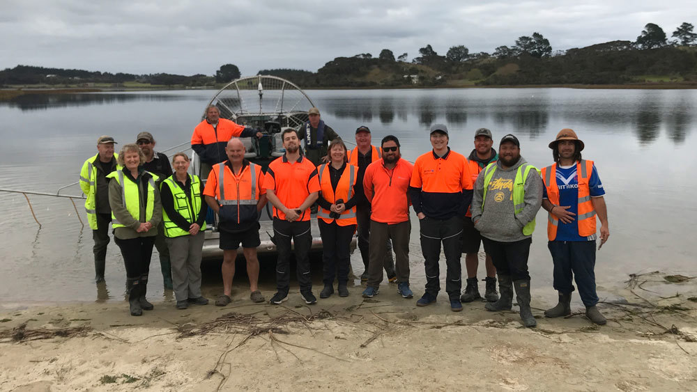 A group of people wearing hi-vis clothing standing by Lake Ngatu, in front of a lake boat.