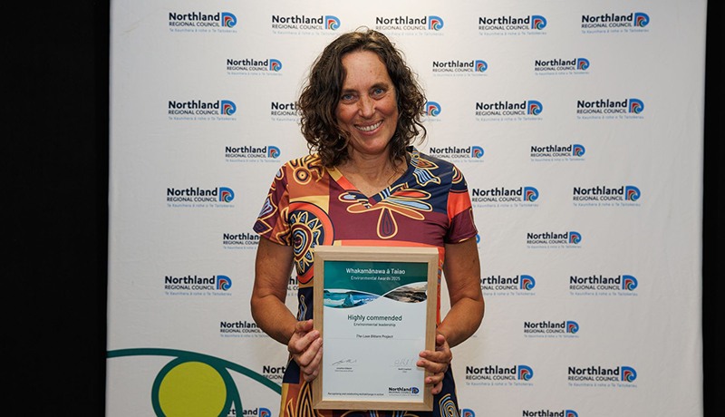 Woman wearing colourful print dress holding a certificate in front of Northland Regional Council logo backdrop. (Photo: Dawn Dutton Photography).