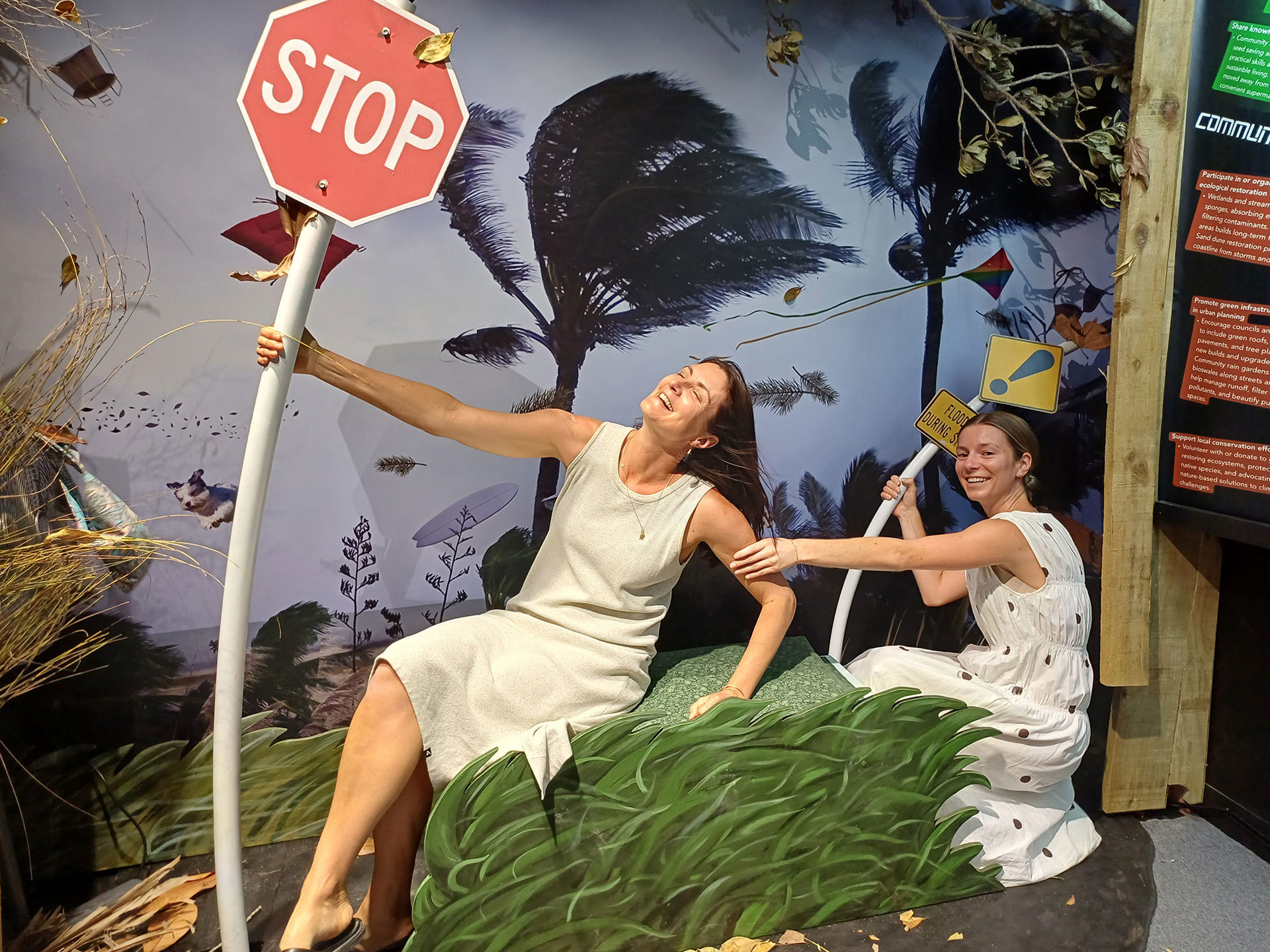 A woman sits in a wind exhibition hanging onto a stop sign pole looking as if she's getting blown away. Another woman sits behind her holding her arm.
