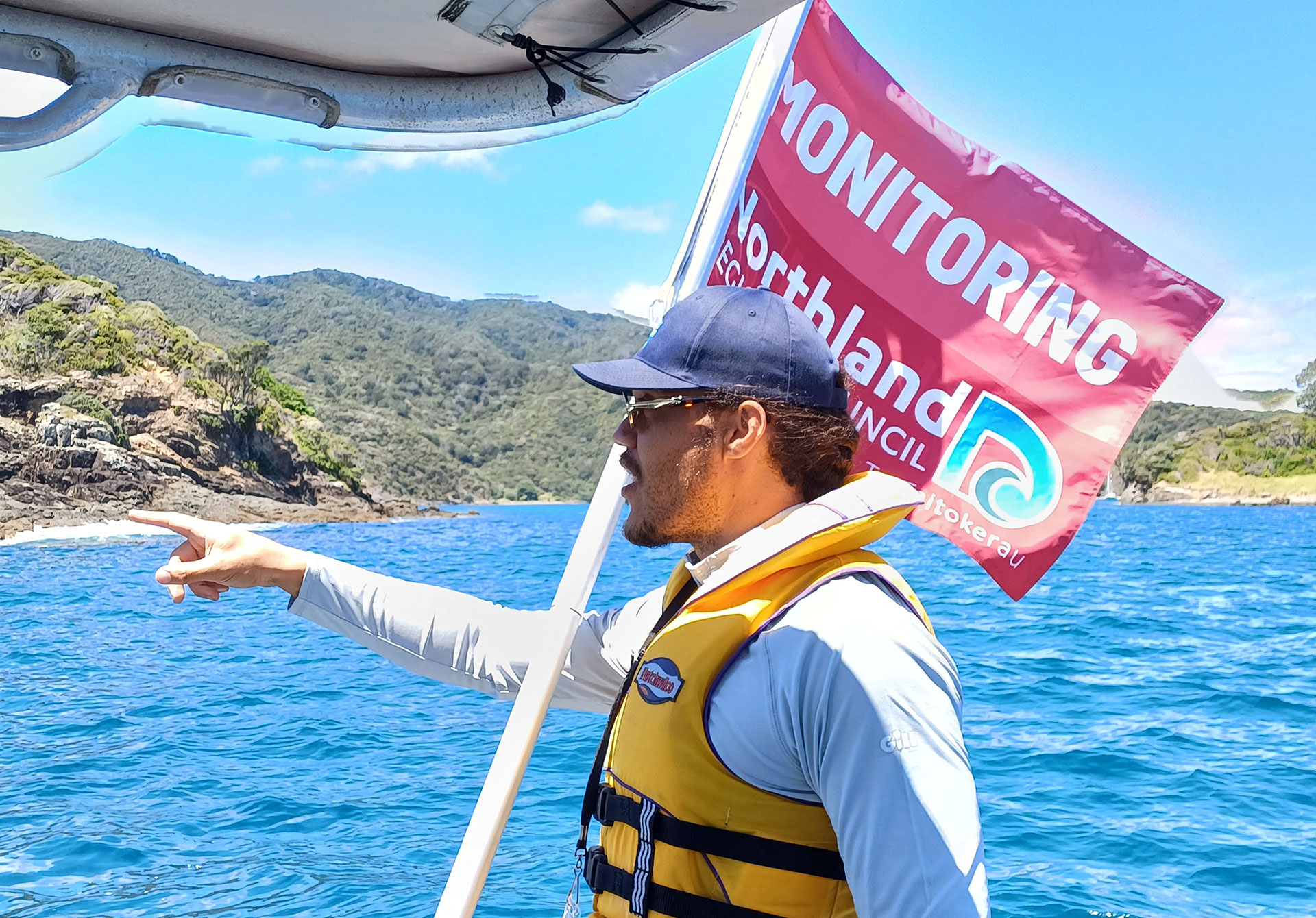 A man on a boat wearing a yellow lifejacket and cap, with red monitoring flag behind stands and points at something.