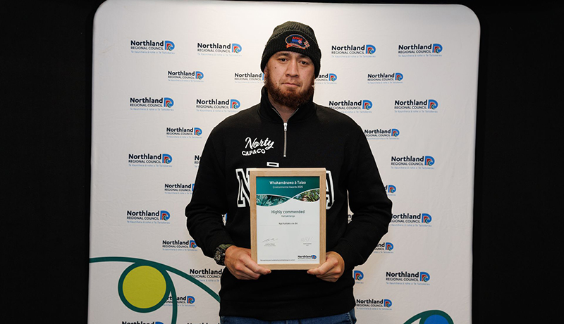 Man wearing a beanie holding a certificate in front of Northland Regional Council logo backdrop. (Photo: Dawn Dutton Photography).