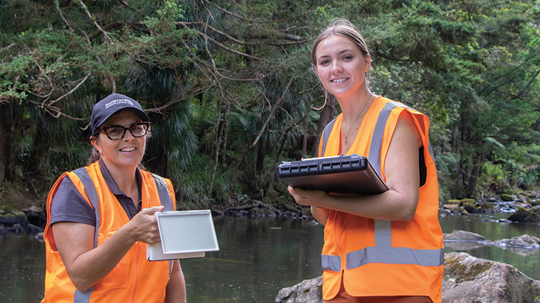 Two woman wearing orange vests holding a clipboard and device with river, rocks and bush in background.