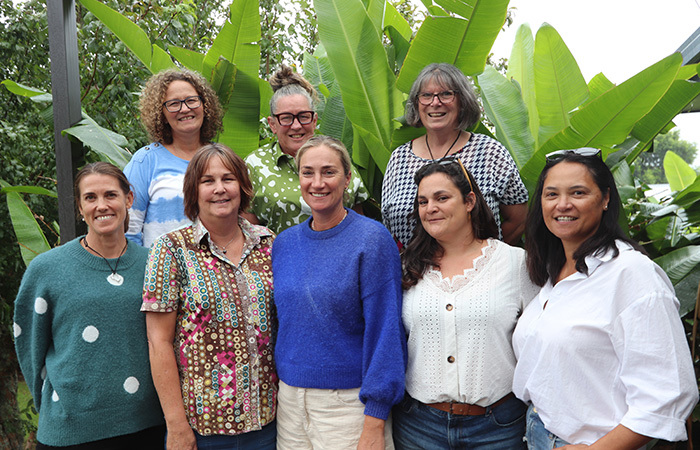 Eight women stand in front of lush green tropical trees
