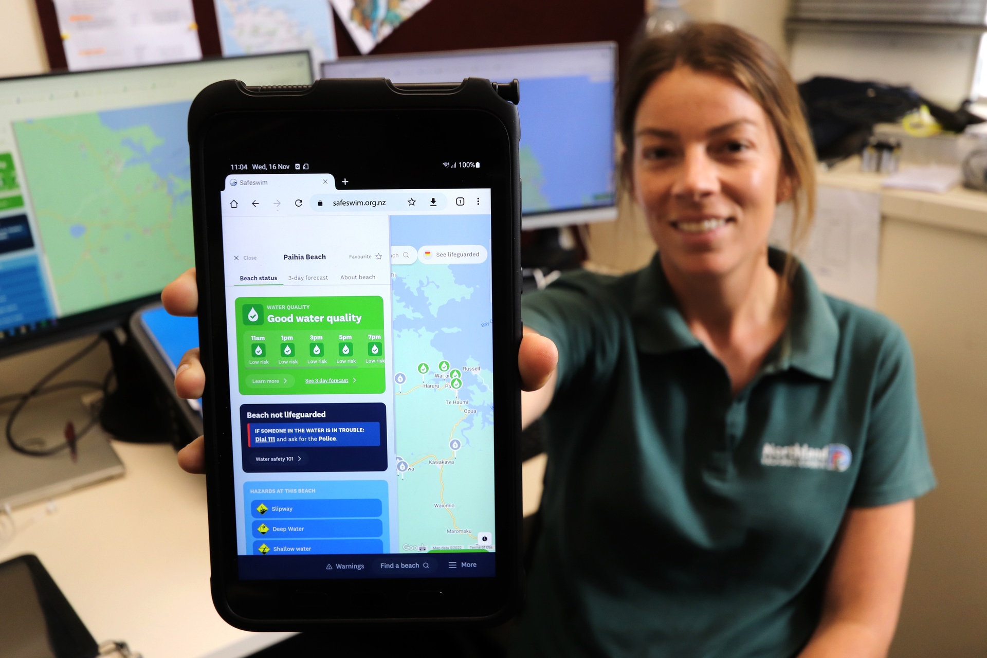 A woman wearing a Northland Regional Council shirt holds a mobile phone with the Safeswim website displayed.