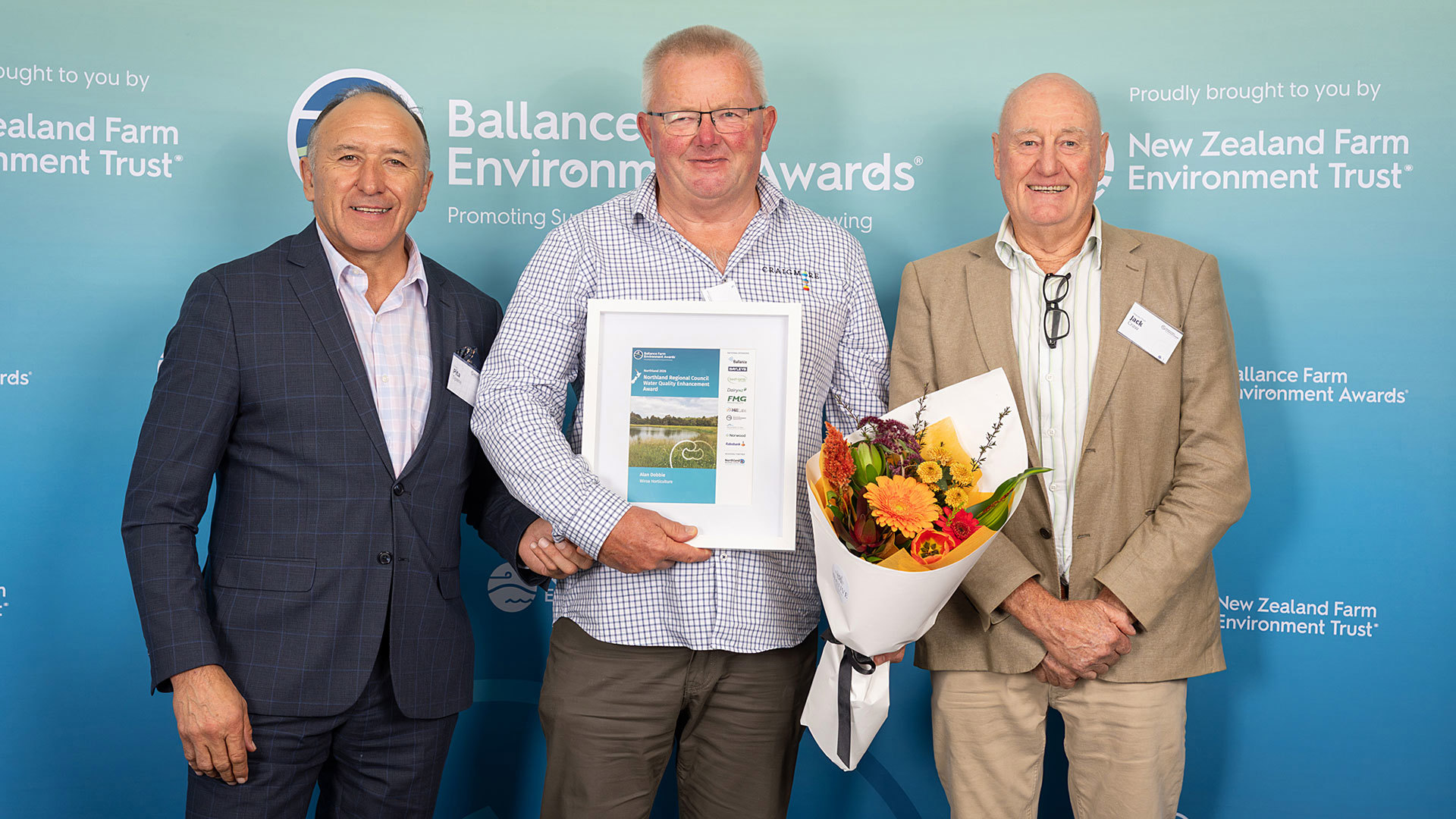 Three men, one holding a certificate and flowers, stand in front of Ballance farm environment awards banner.