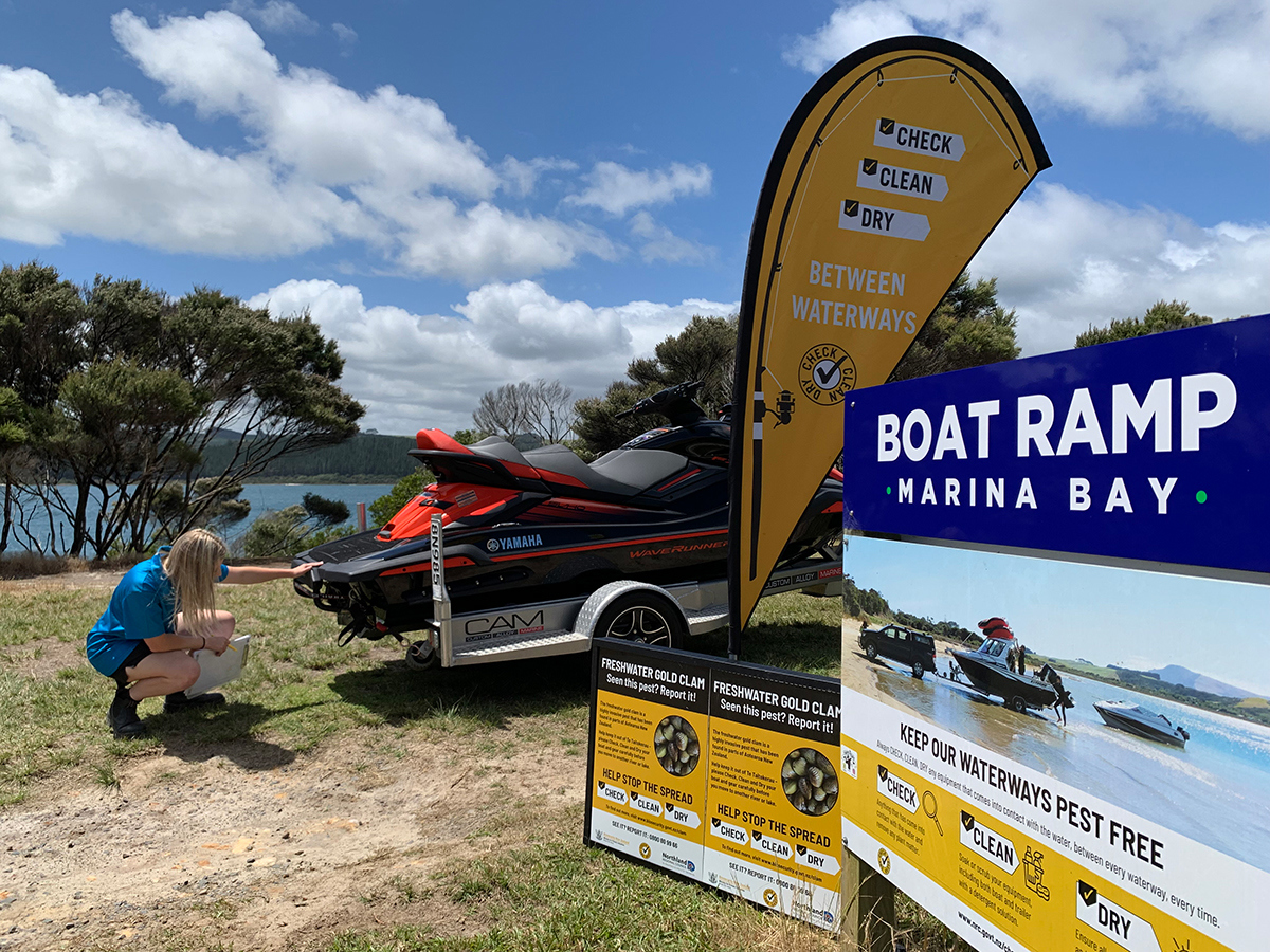 A woman inspects the stern of a jet ski on a trailer with check, clean, dry and freshwater invasive clam signs.