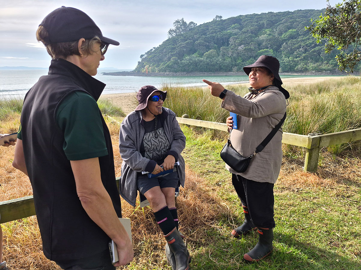 Three women wearing hats, and two wearing gumboots standing or sitting near dune plantings with the coast in the background.