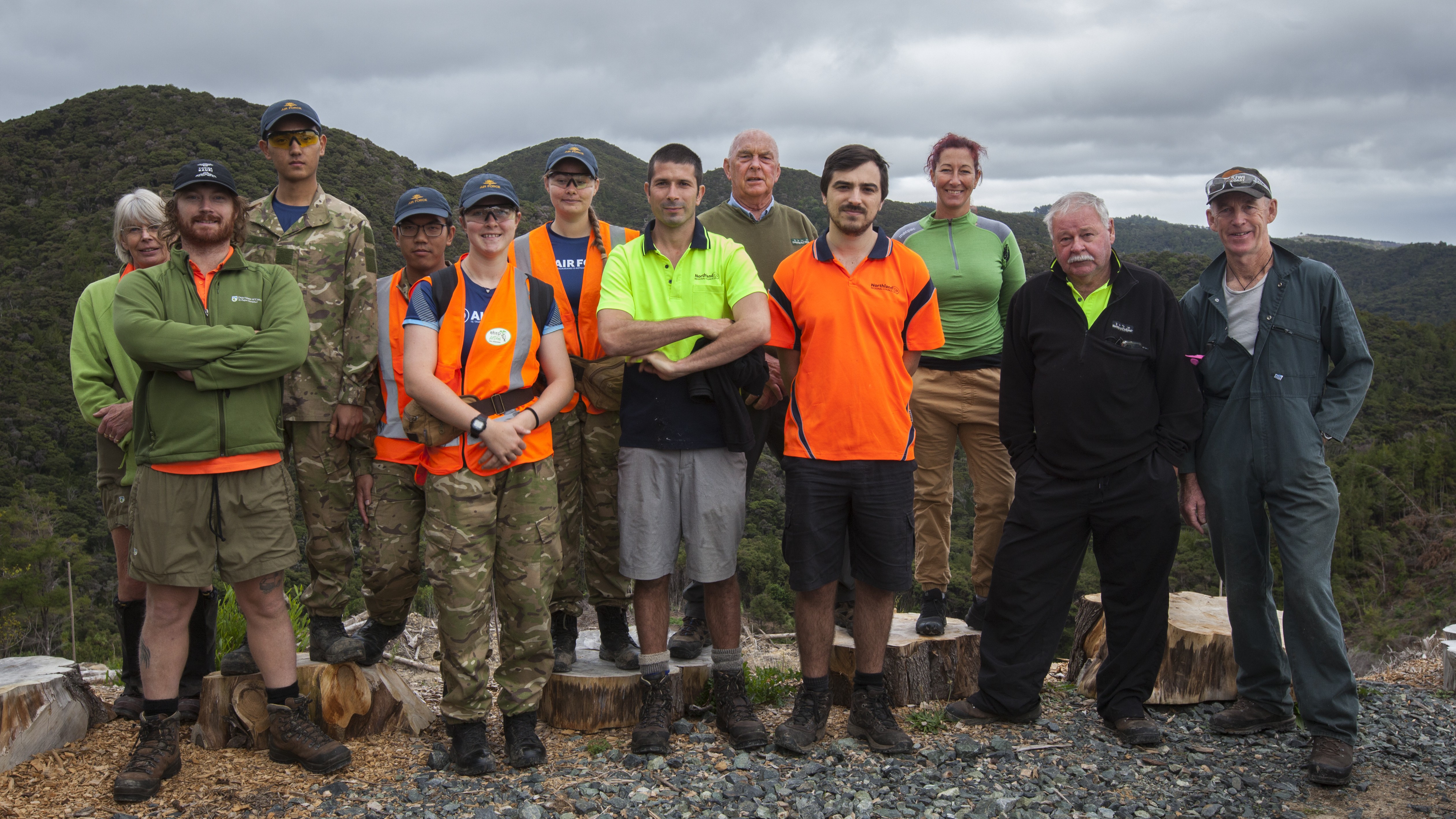 A group of volunteers and project partners stand on a hilltop overlooking bush-covered valleys. Several wear high-visibility work clothing and outdoor gear, with cut tree stumps and native forest visible in the foreground and background.