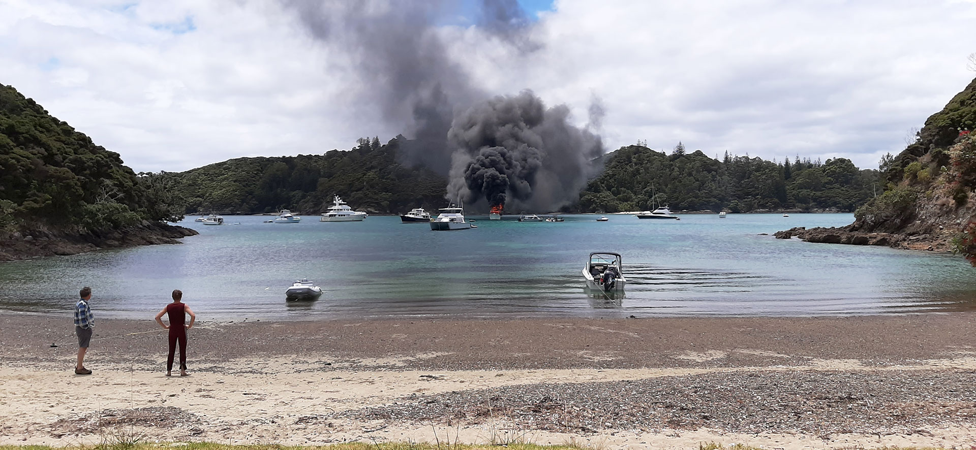 Clouds of black smoke above a boat on fire in a bay, with other boats nearby. Two men stand onshore watching.